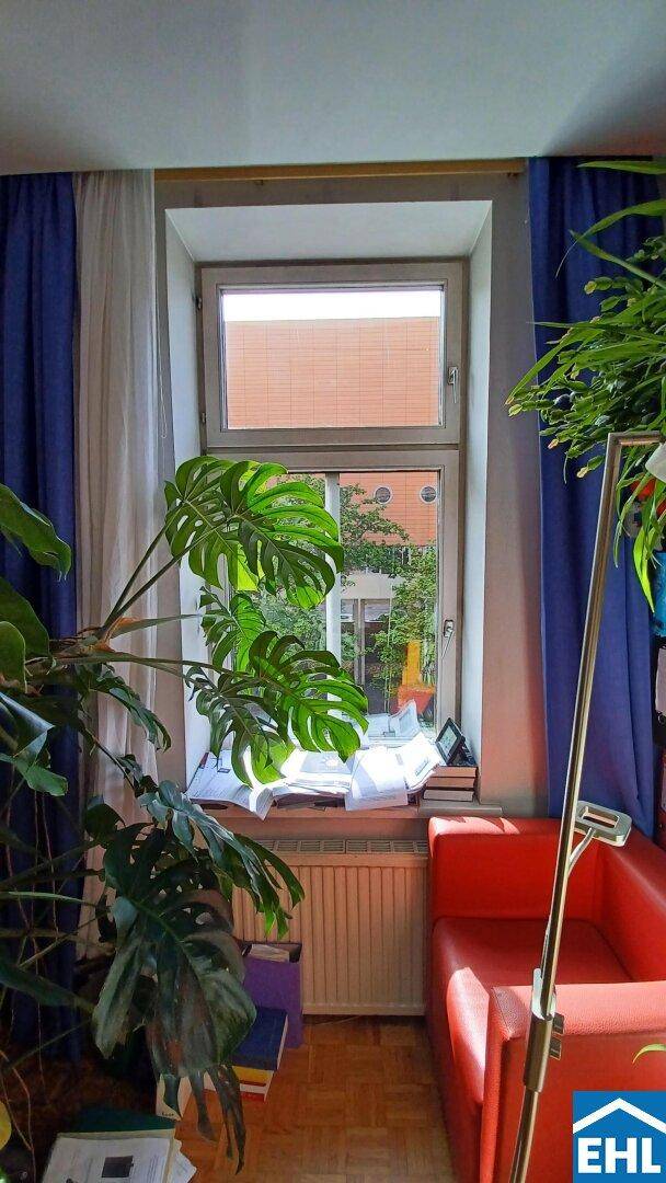 Gemütliches Wohnzimmer mit großem Fenster, vielen Pflanzen und einem roten Sofa.