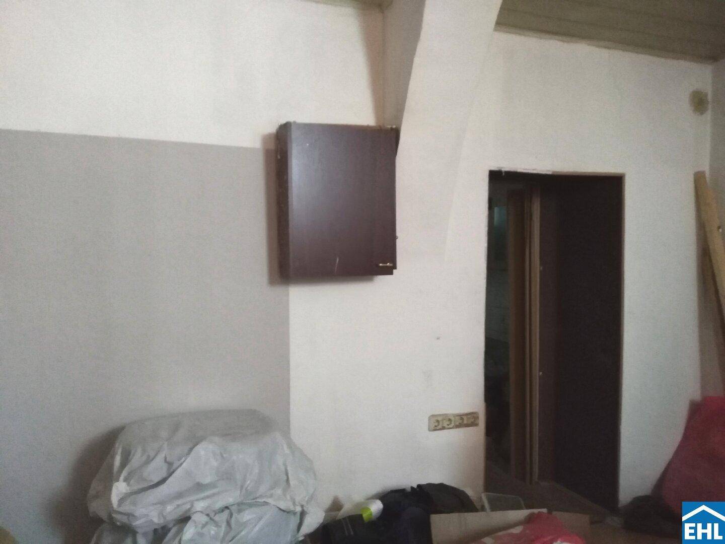 Eine Innenansicht mit einer Wand, einem Wandschrank und einem Türrahmen, der in einen weiteren Raum führt.