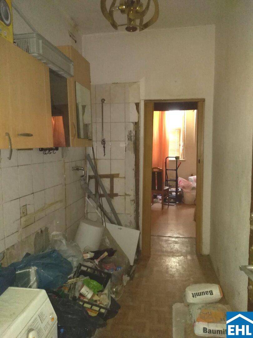 Ein alter Küchenbereich mit gefliesten Wänden und Hängeschränken, der in einen weiteren Raum führt.