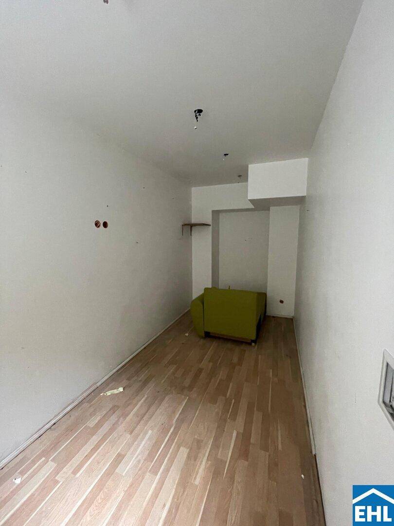 Kleiner, leerer Raum mit weißen Wänden, Laminatboden und einem einzelnen grünen Sessel.