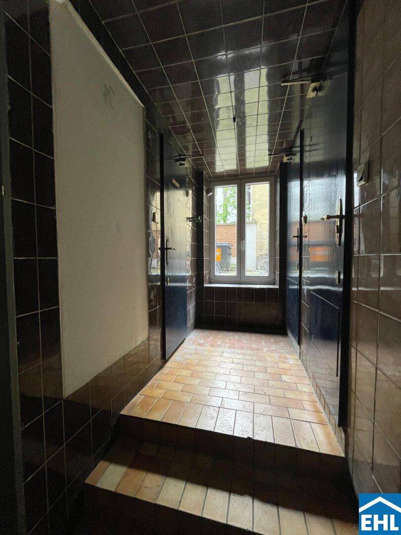 Raum mit dunkelbraunen Fliesen an Wänden und Boden, mehreren Türen und einem Fenster am Ende.