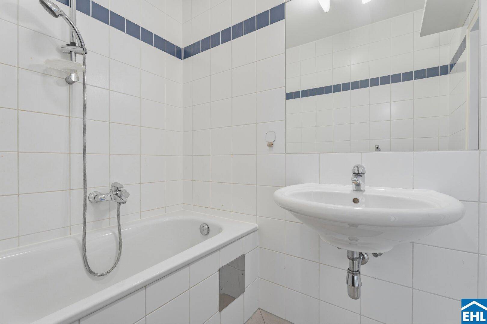 Badezimmer mit Badewanne, Waschbecken und weißen Fliesen mit blauer Bordüre.