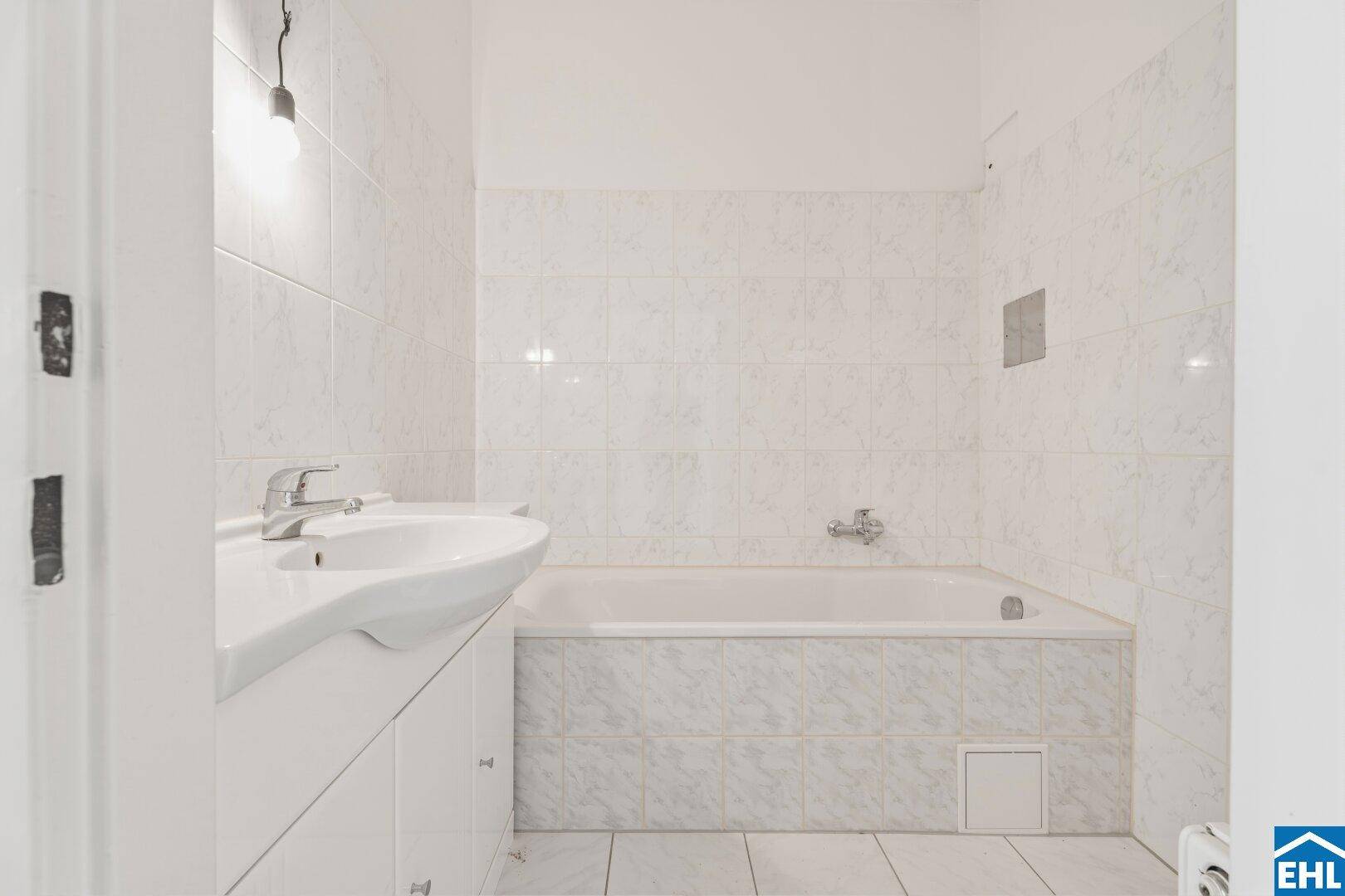 Helles Badezimmer mit Badewanne und Waschbecken, umgeben von weißen Wandfliesen mit Muster.
