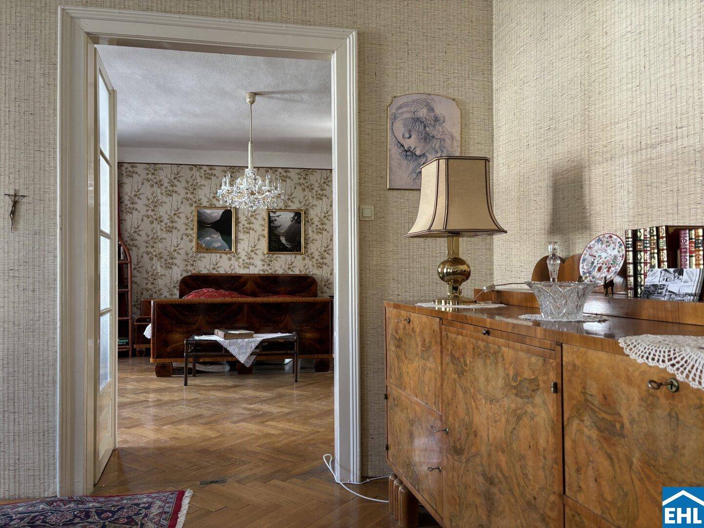 Heller Flur mit Kommode und Blick in ein Schlafzimmer mit Kronleuchter und Parkettboden.
