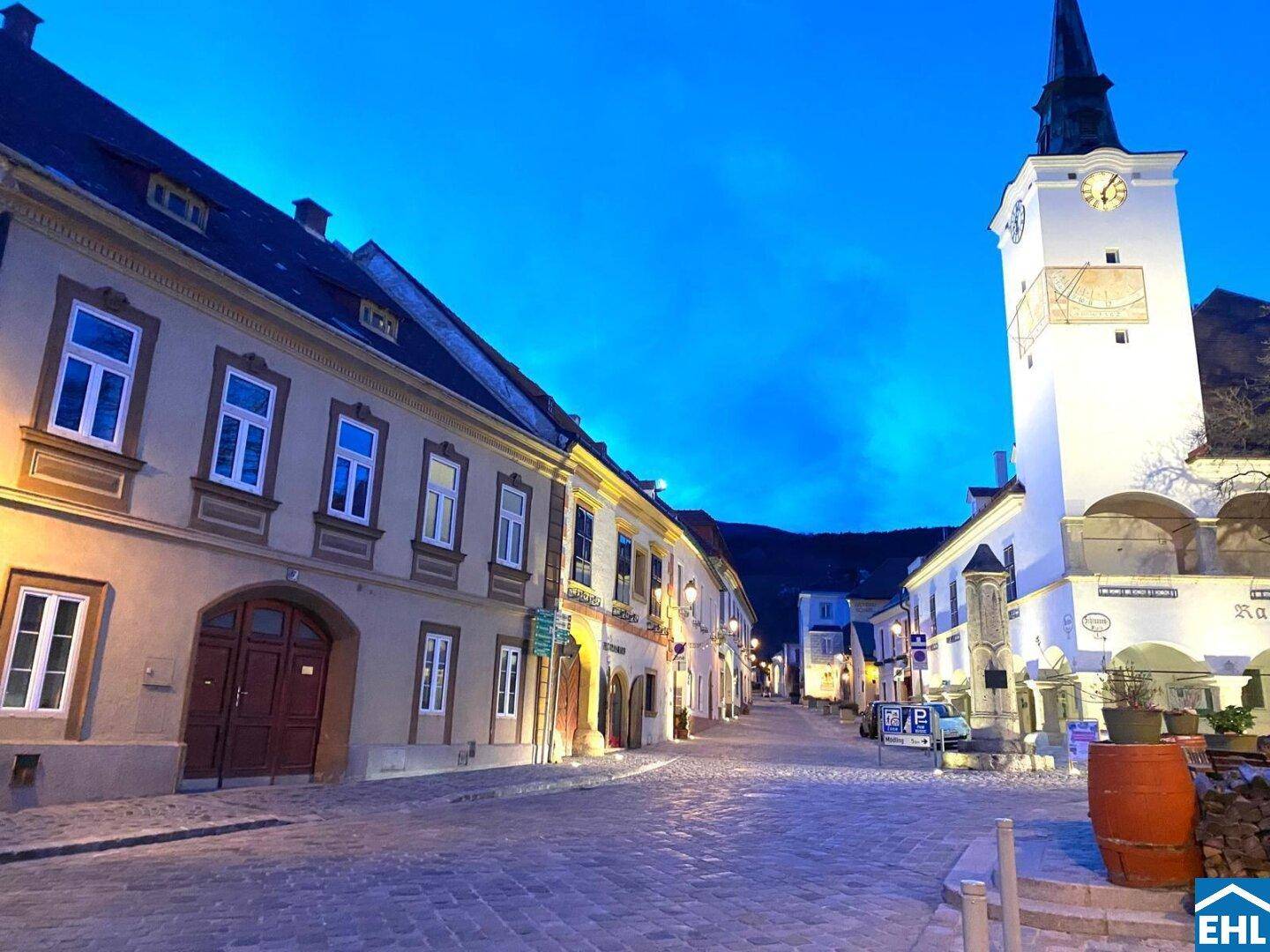 Malerische Altstadtgasse bei Dämmerung mit beleuchteten Gebäuden und einem markanten Kirchturm.