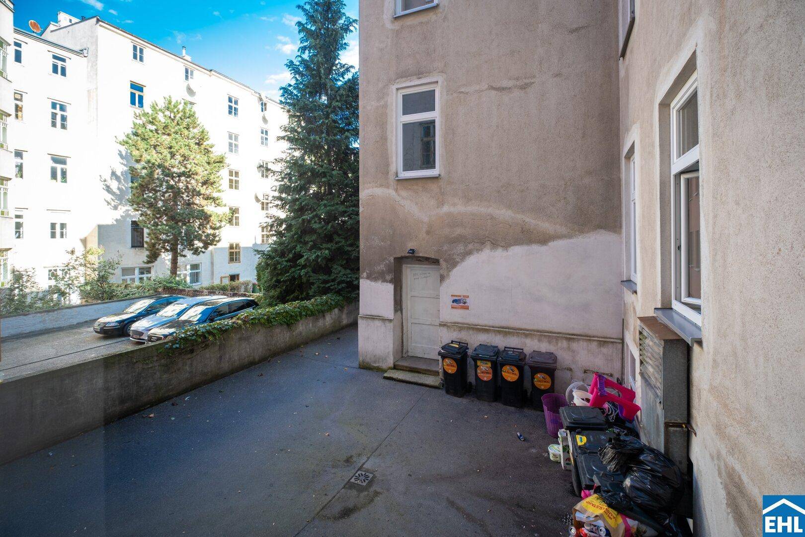 Hinterhofbereich mit Mülltonnen, Parkplätzen und Bäumen, umgeben von Gebäuden.