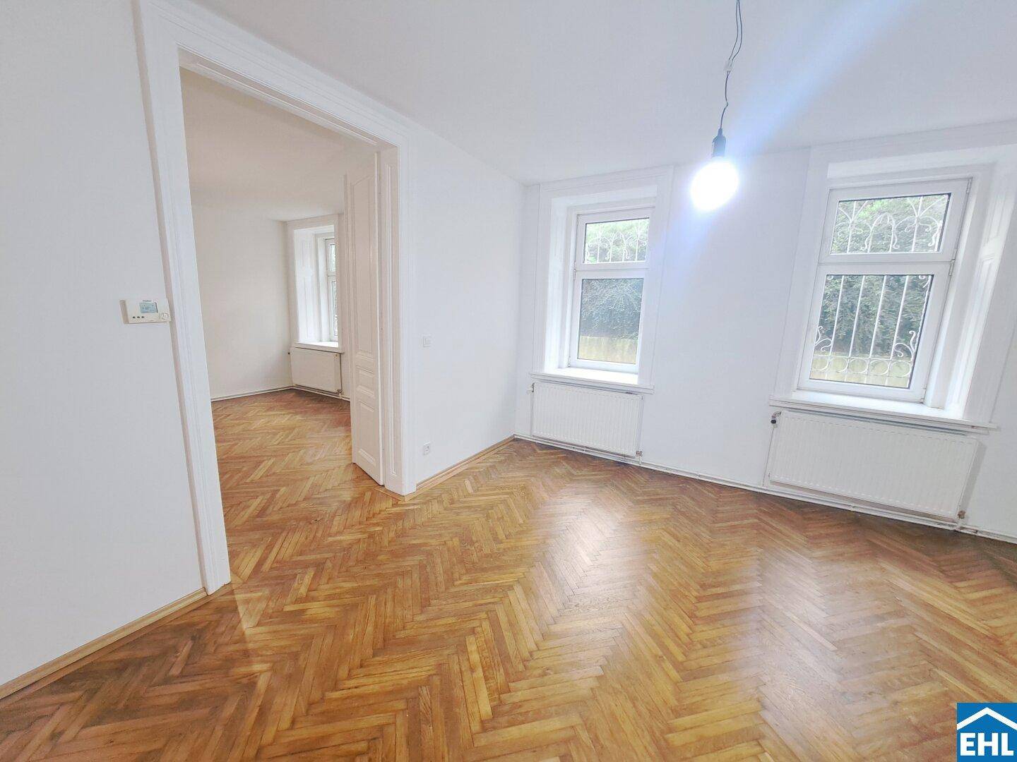Lichtdurchflutetes Zimmer mit Fischgrätparkett, zwei Fenstern und Blick in einen angrenzenden Raum.