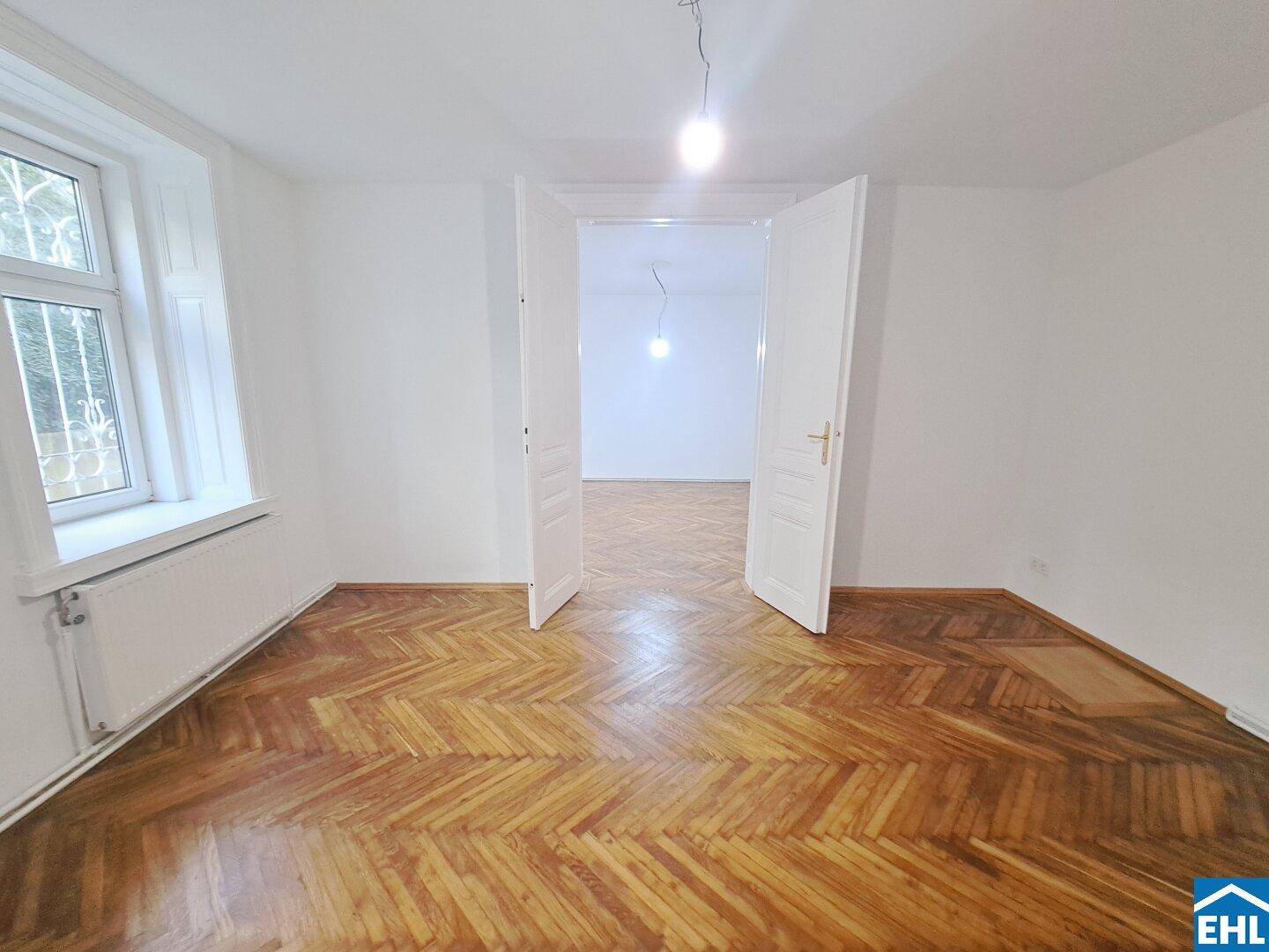 Geräumiges Zimmer mit klassischem Fischgrätparkett und hohen Doppeltüren zu einem weiteren Raum.