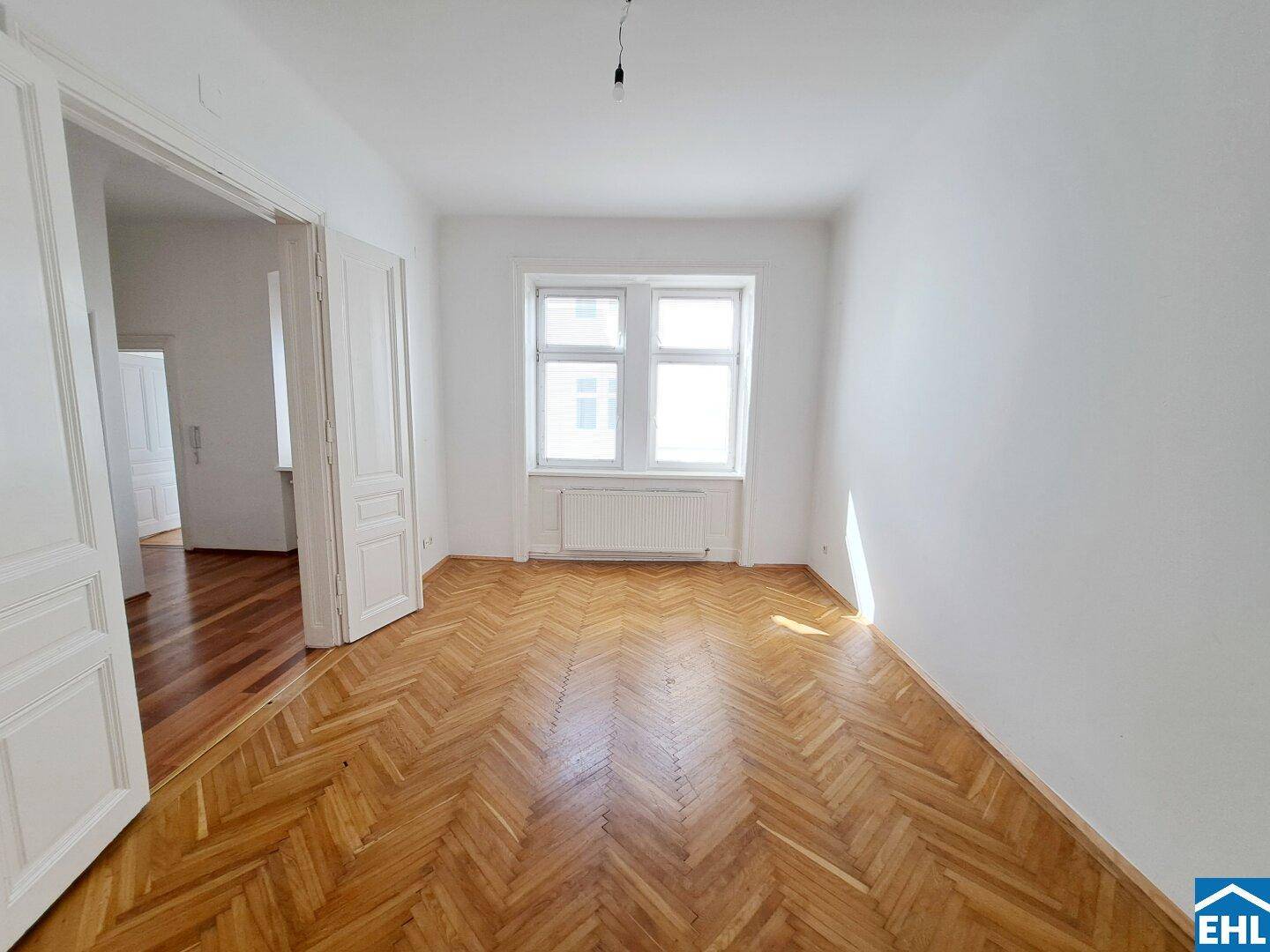Lichtdurchflutetes Zimmer mit Fischgrätparkett und zwei großen Fenstern.