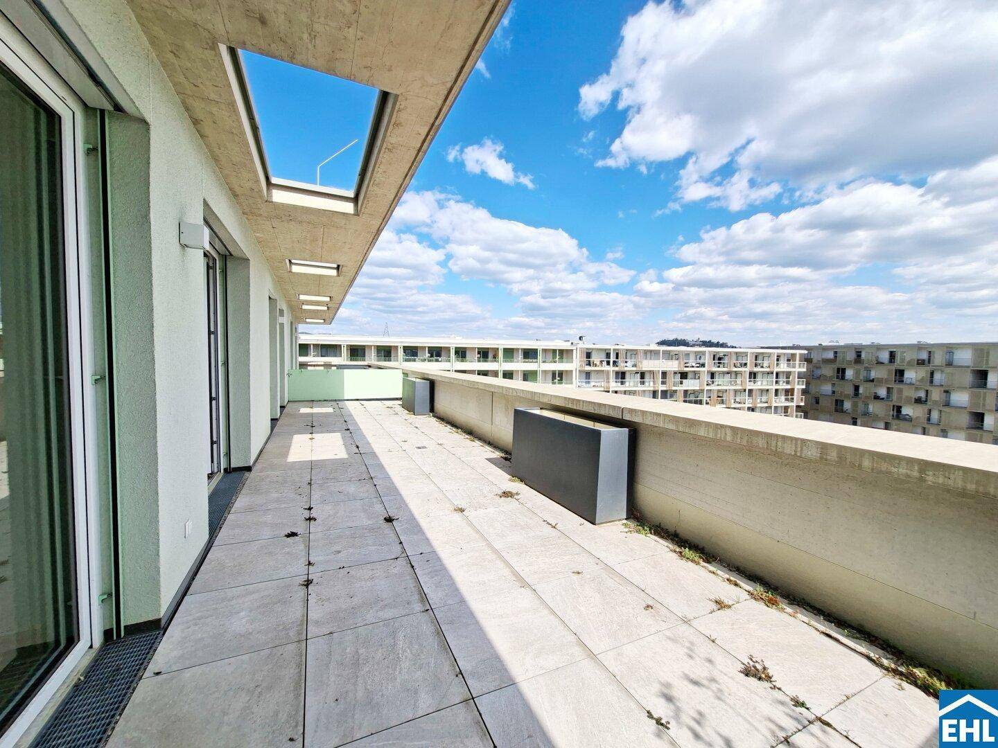 Lange Dachterrasse mit Fliesenboden, umgeben von Gebäuden und mit Blick in den Himmel.