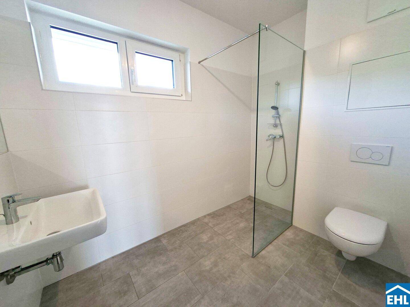 Modernes Badezimmer mit ebenerdiger Dusche, wandhängendem WC und Fenster für Helligkeit.