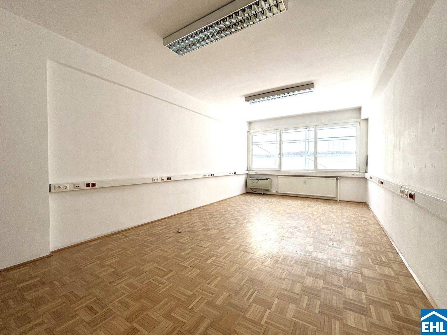 Geräumiges Büro mit Parkettboden und breiter Fensterfront, bietet viel Tageslicht.