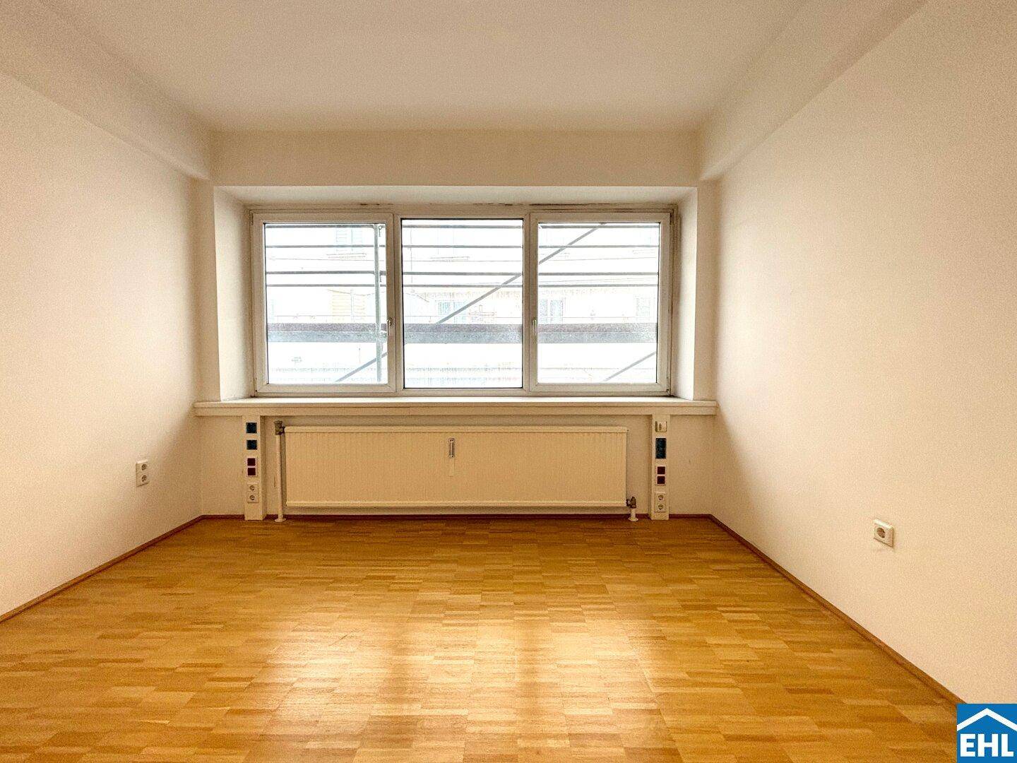 Heller, leerer Raum mit Parkettboden und breitem Fenster mit Heizkörper.