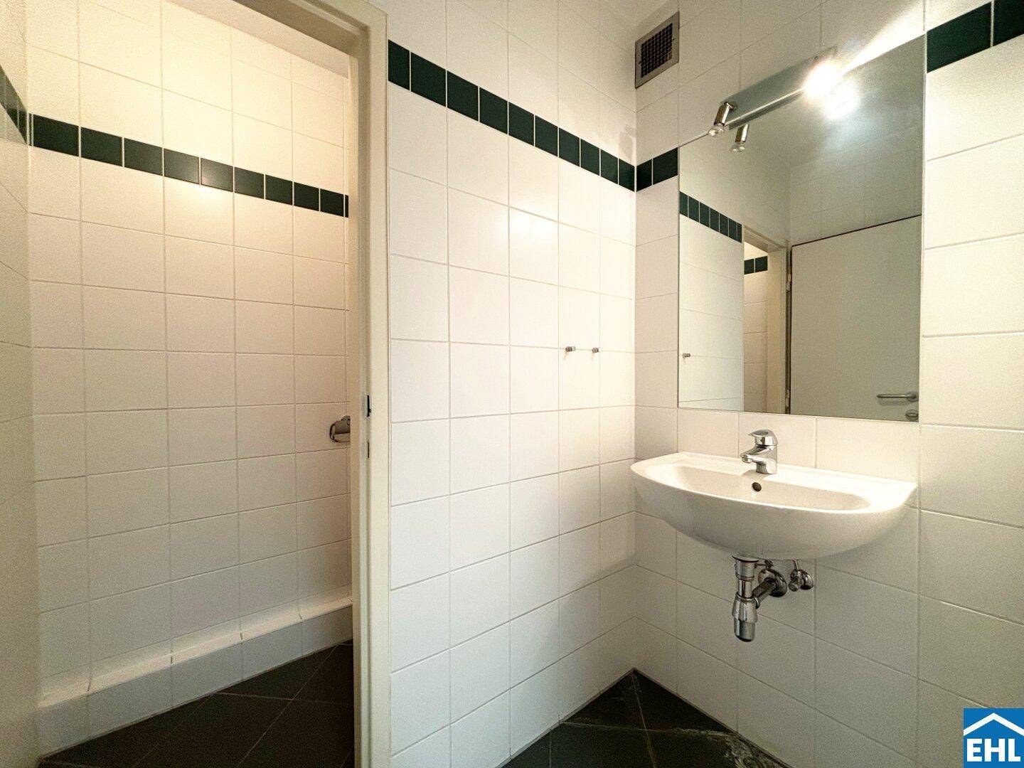 Badezimmer mit weißen Fliesen, einem Waschbecken und einer Duschvorrichtung.