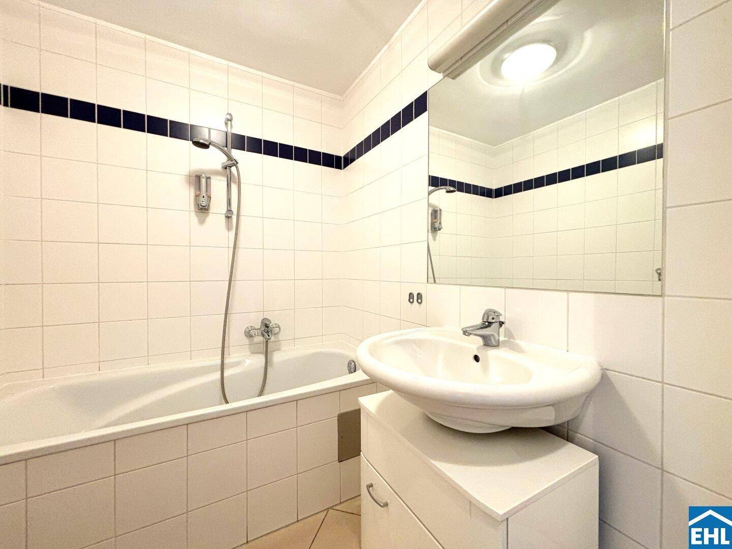 Badezimmer mit Badewanne, Waschbecken und weißen Fliesen, akzentuiert durch eine blaue Bordüre.