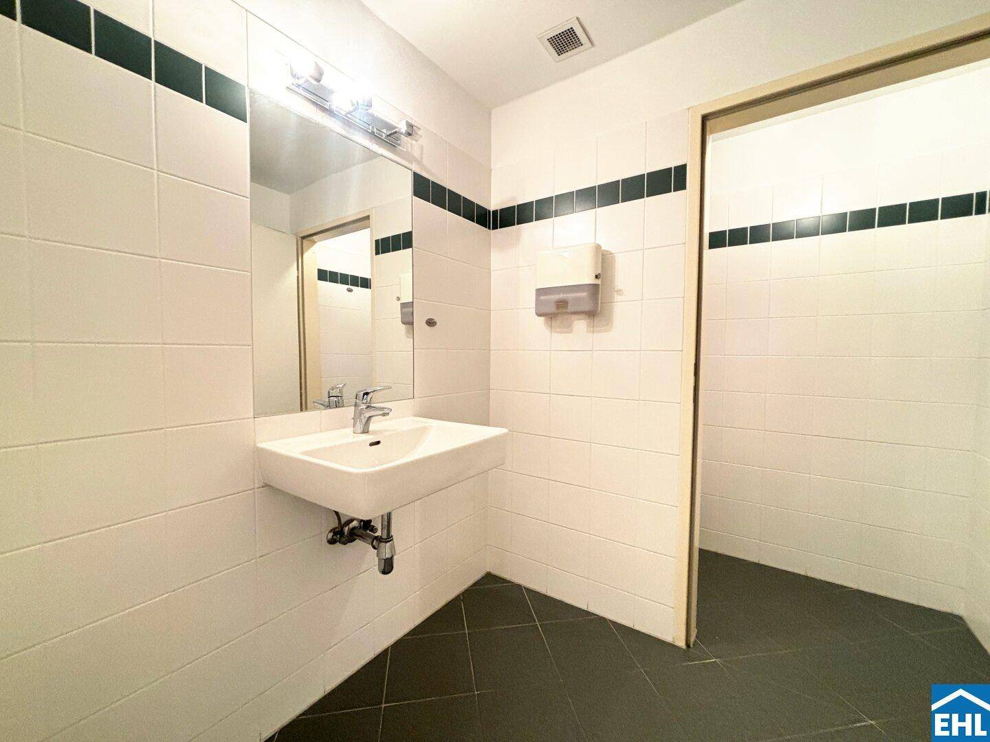 Einfaches WC mit weiß gefliesten Wänden und dunklem Boden, ausgestattet mit einem einzelnen Waschbecken.