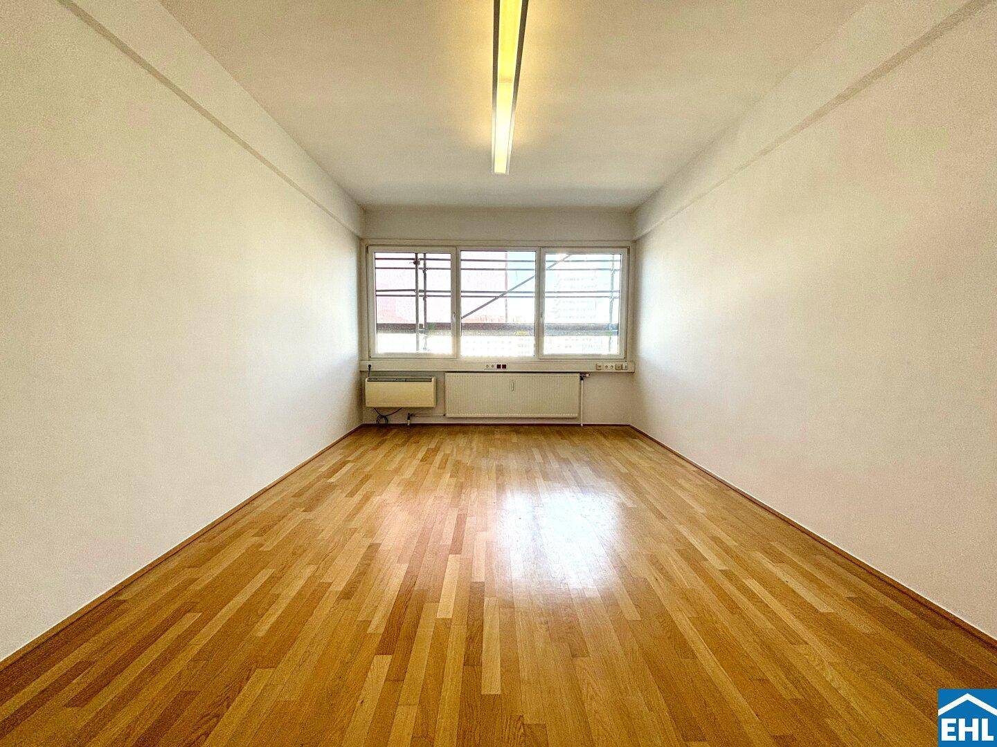 Geräumiger, leerer Raum mit hellem Holzparkett und breiter Fensterfront für viel Tageslicht.