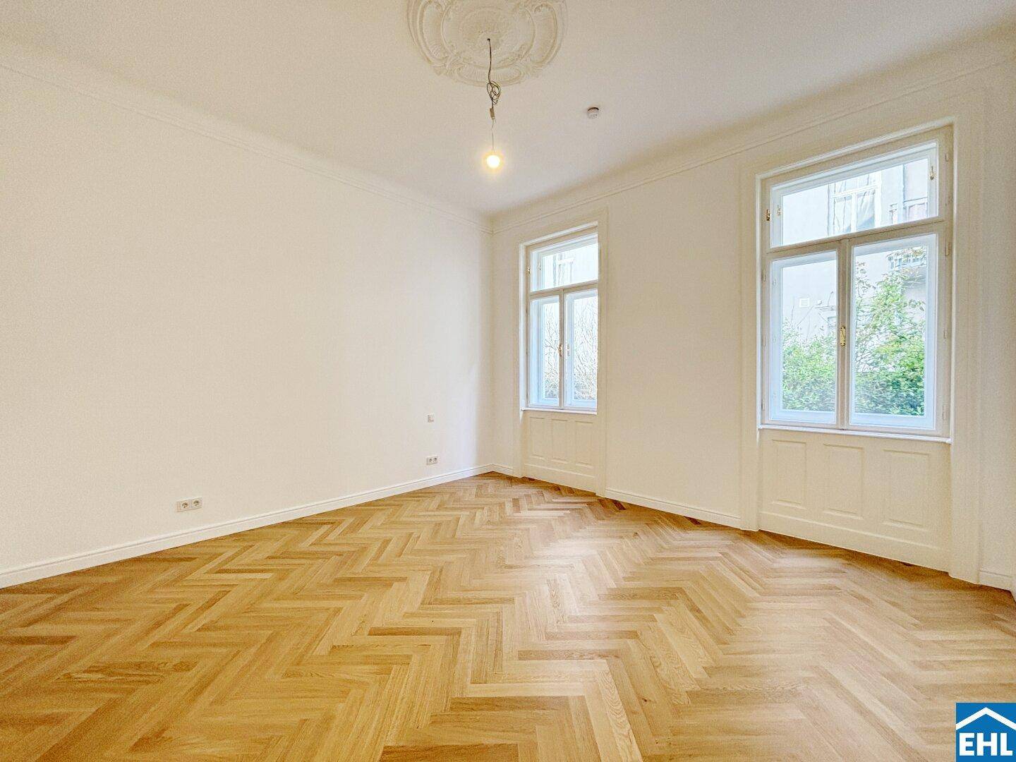 Lichtdurchfluteter Raum mit elegantem Fischgrätparkett und zwei hohen Fenstern.