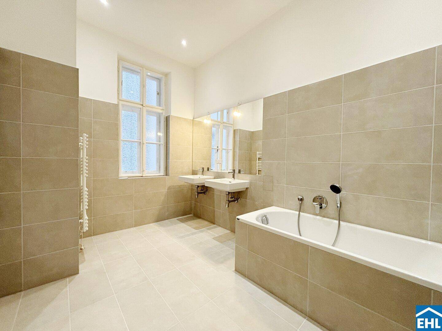Großes Badezimmer mit Doppelwaschbecken, Badewanne und beigefarbenen Fliesen an Wänden und Boden.