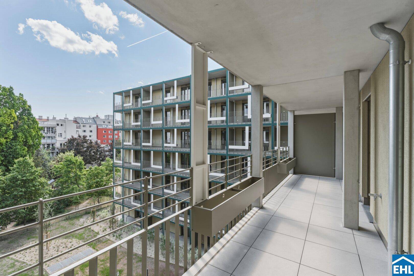 Langer Balkon mit Fliesenboden und Blick auf die moderne Wohnanlage und Grünflächen.