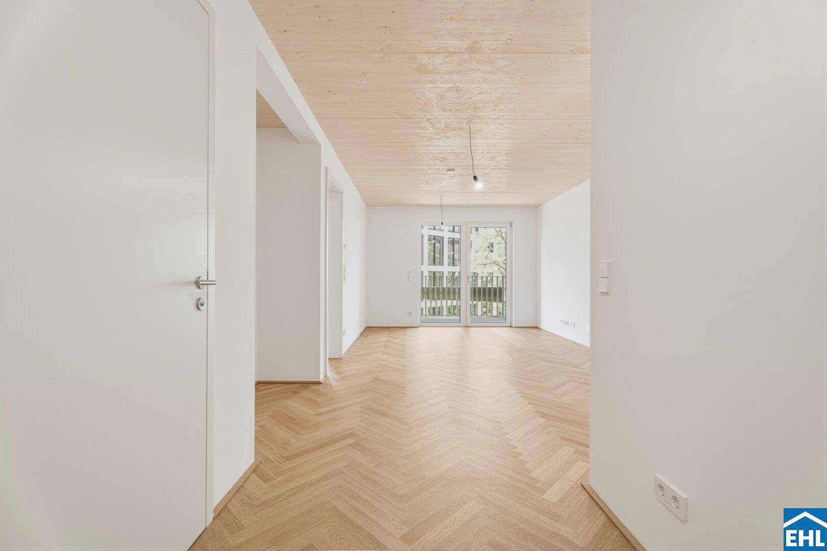 Blick vom Eingangsbereich in ein helles Zimmer mit Fischgrätparkett und Balkonzugang.