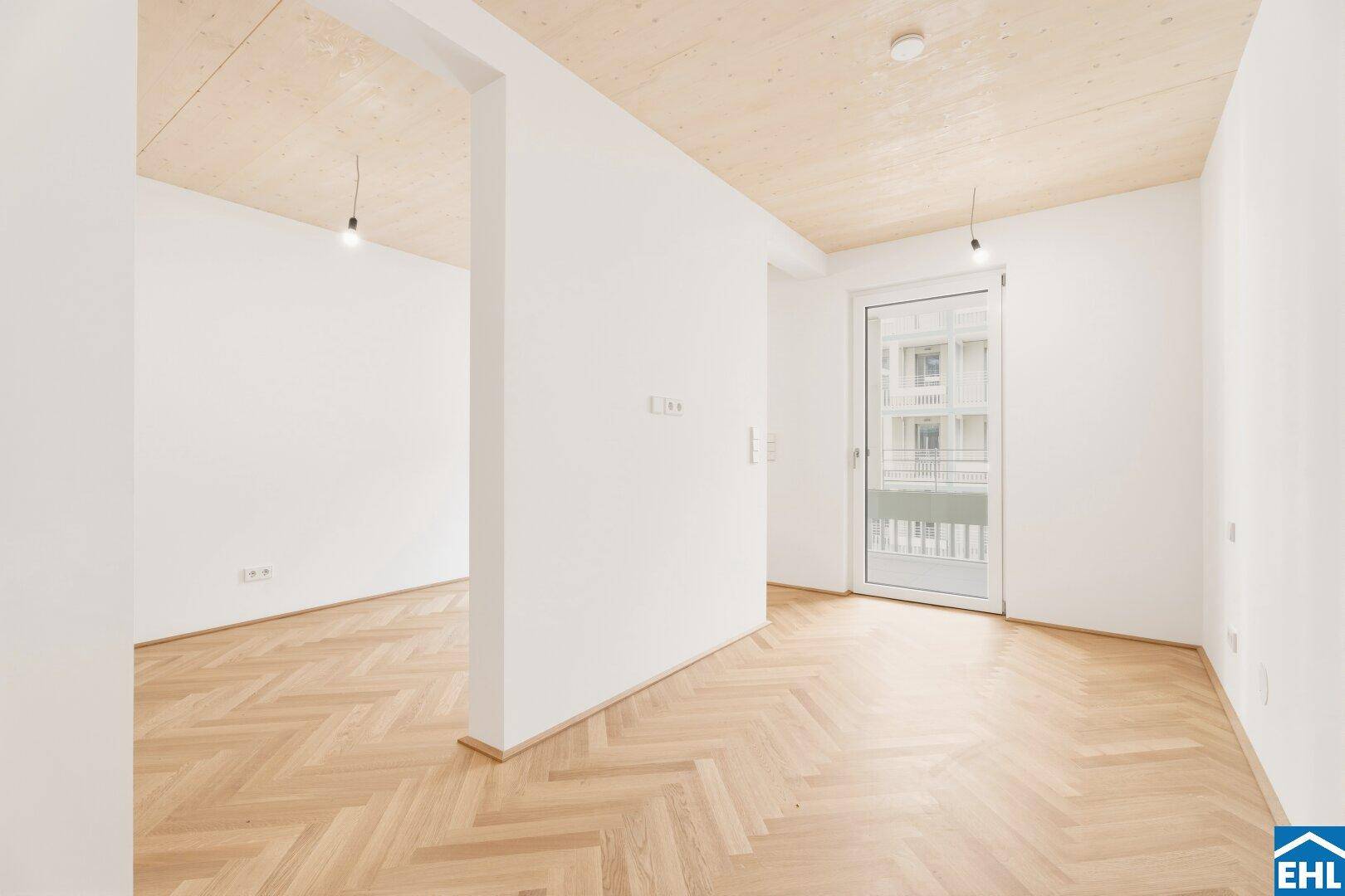 Leeres Zimmer mit Fischgrätparkett, Holzbalkendecke und viel Tageslicht durch die Tür.