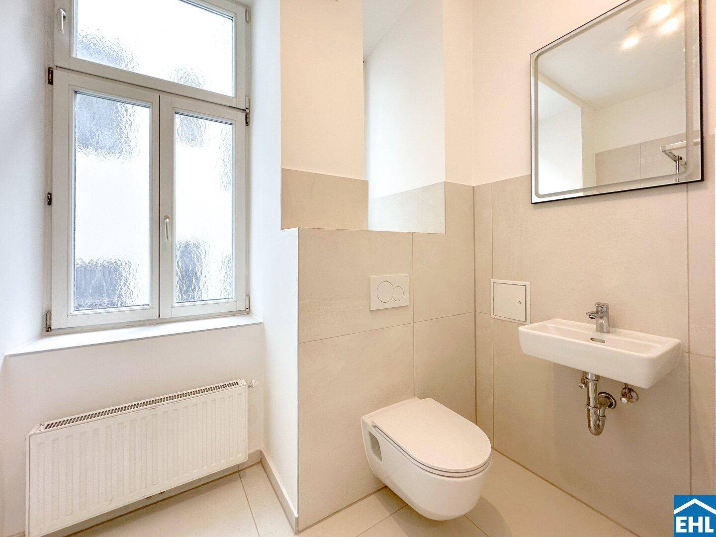Separates WC mit wandhängender Toilette, kleinem Waschbecken und Fenster.