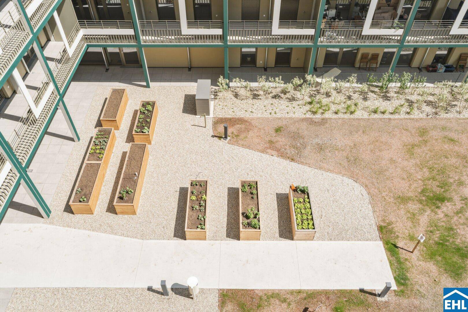 Gemeinschaftsgarten mit Hochbeeten und Kiesflächen, umgeben von moderner Wohnbebauung.