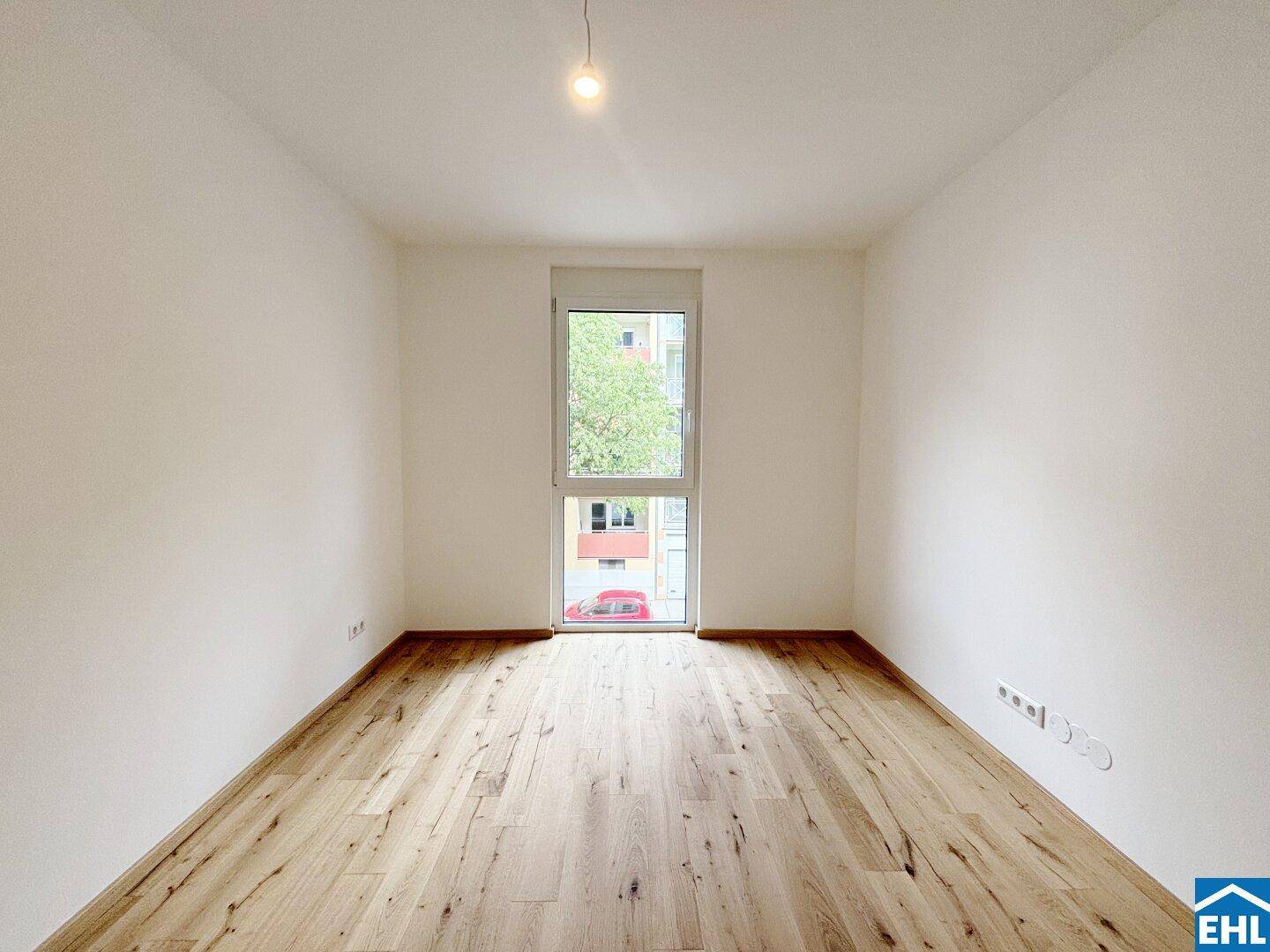 Leeres Schlafzimmer mit hellem Holzboden und einem großen Fenster, das viel Tageslicht hereinlässt.