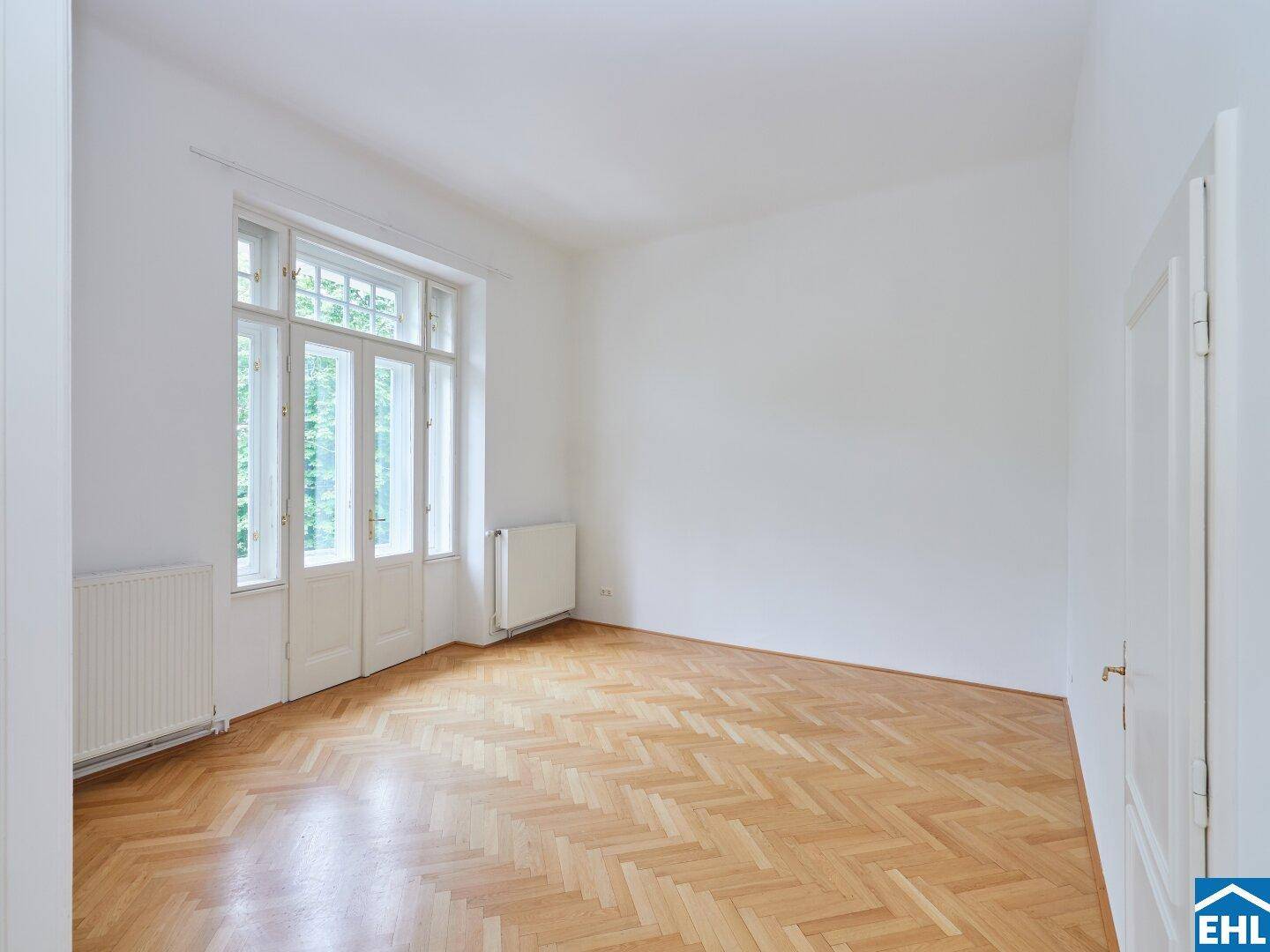 Geräumiger Raum mit Fischgrätparkett und großen Fenstertüren, die viel Licht hereinlassen.