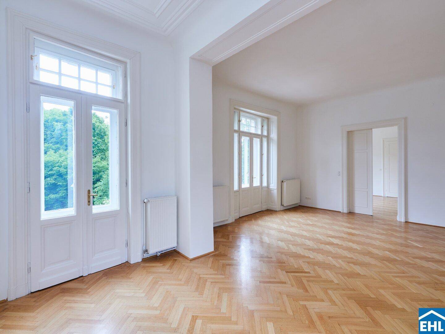 Heller Wohnbereich mit Fischgrätparkett und hohen Fenstertüren, die den Zugang nach draußen ermöglichen.