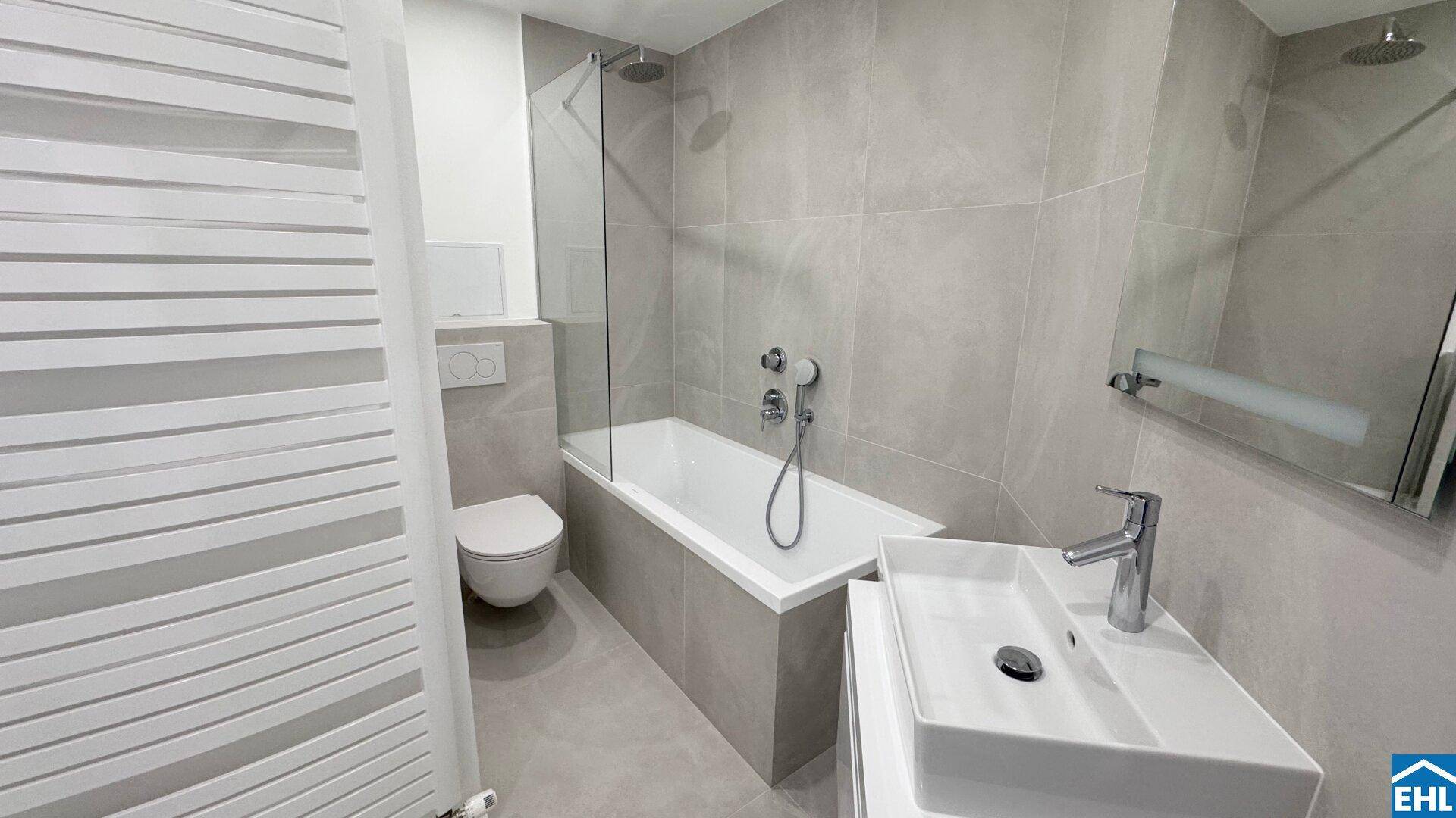 Modernes Badezimmer mit Badewanne, Dusche, WC und grau gefliesten Wänden und Boden.