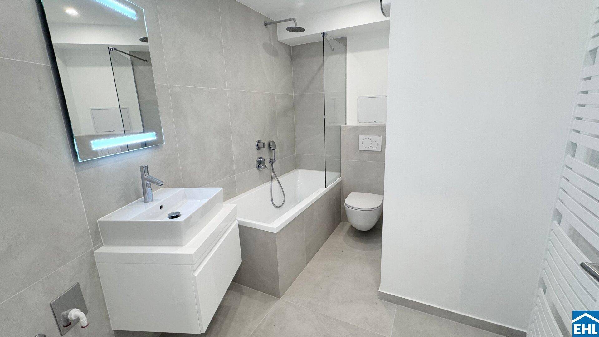 Modernes Badezimmer mit grauen Fliesen, einer Badewanne mit Duschfunktion und einem wandhängenden WC.
