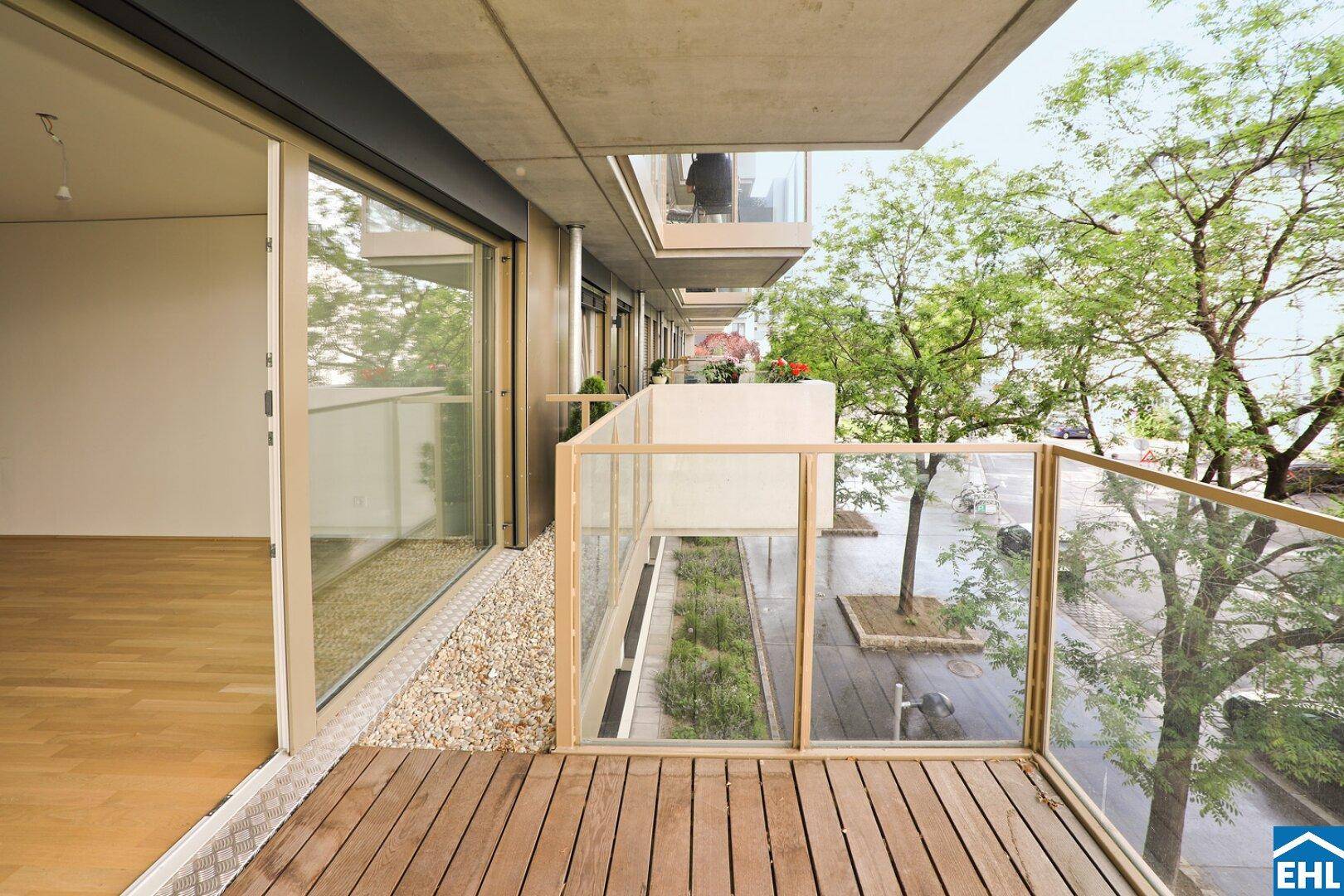 Geräumiger Balkon mit Holzdeck und Glasgeländer, mit Blick auf den begrünten Innenhof.