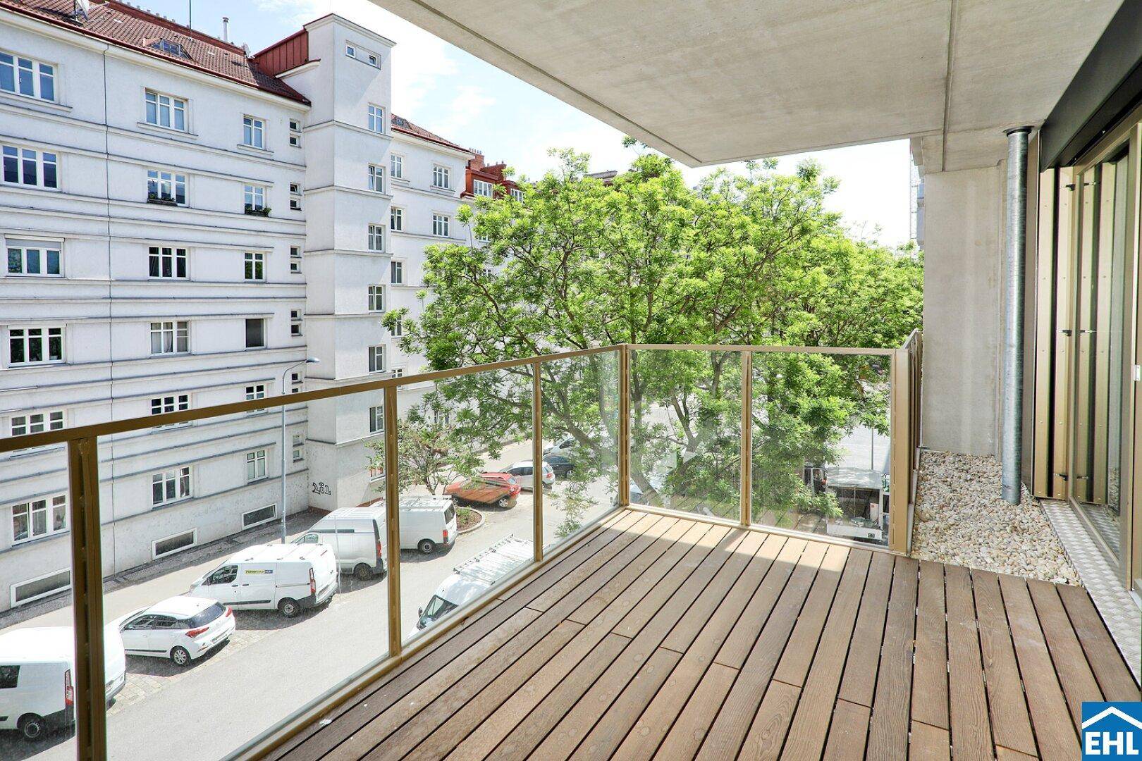 Geräumiger Balkon mit Holzboden und Glasgeländer, bietet Blick auf die Straße und Bäume.