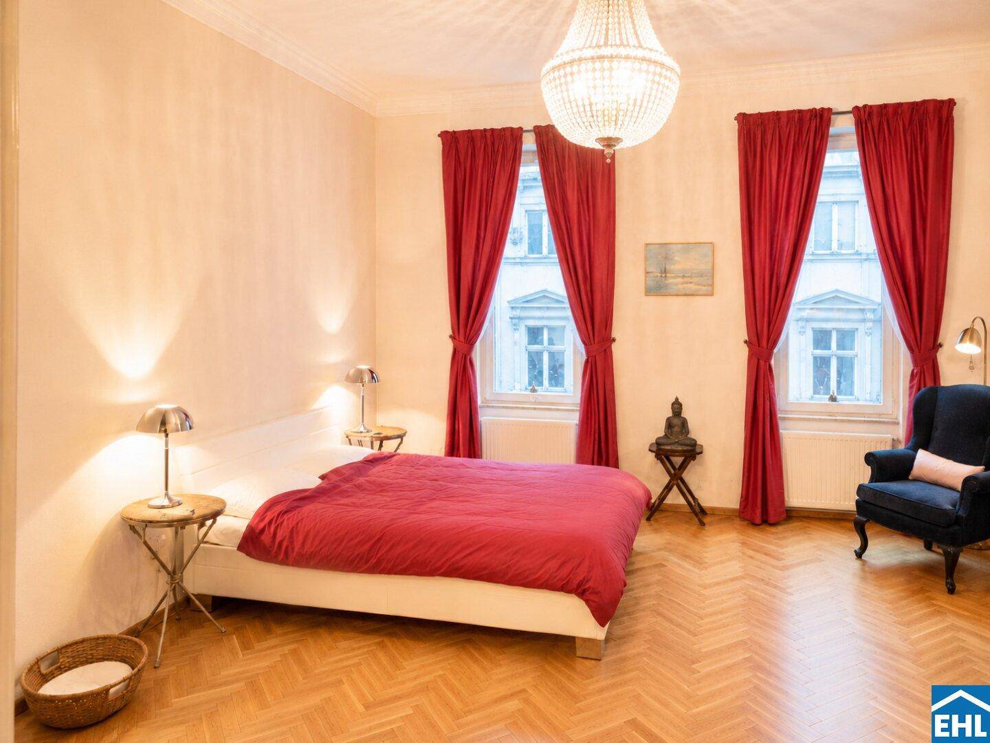 Geräumiges Schlafzimmer mit roter Bettwäsche, Fischgrätparkett und einem eleganten Kronleuchter.