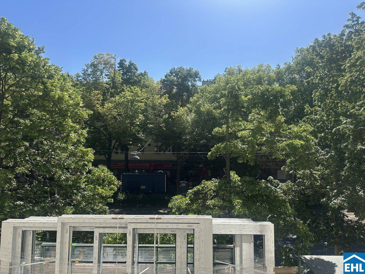 Blick über grüne Baumwipfel mit gestapelten neuen Fensterrahmen im Vordergrund unter blauem Himmel.