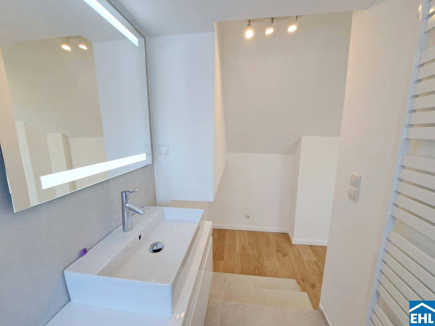 Modernes Badezimmer mit Einzelwaschbecken, beleuchtetem Spiegel und Zugang zu einem weiteren Bereich.