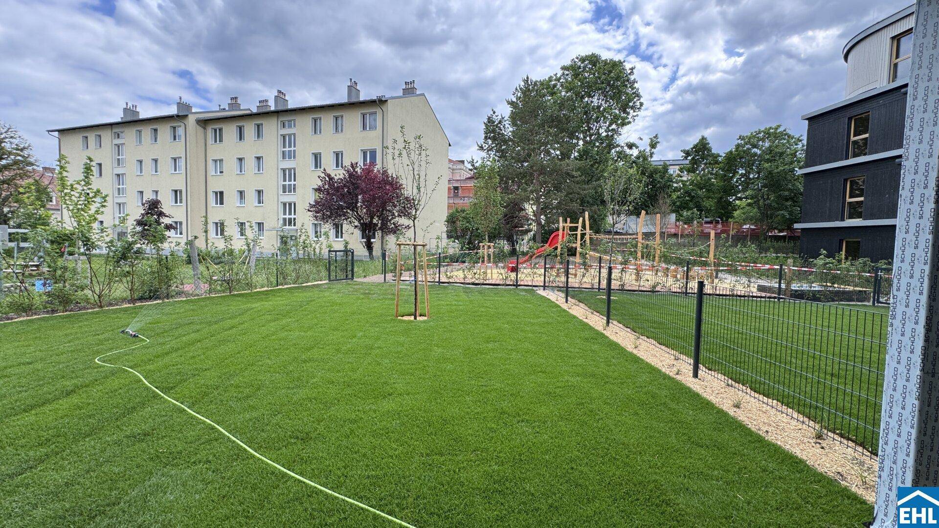 Gepflegte Rasenfläche mit Kinderspielplatz im Hintergrund und Wohngebäuden.