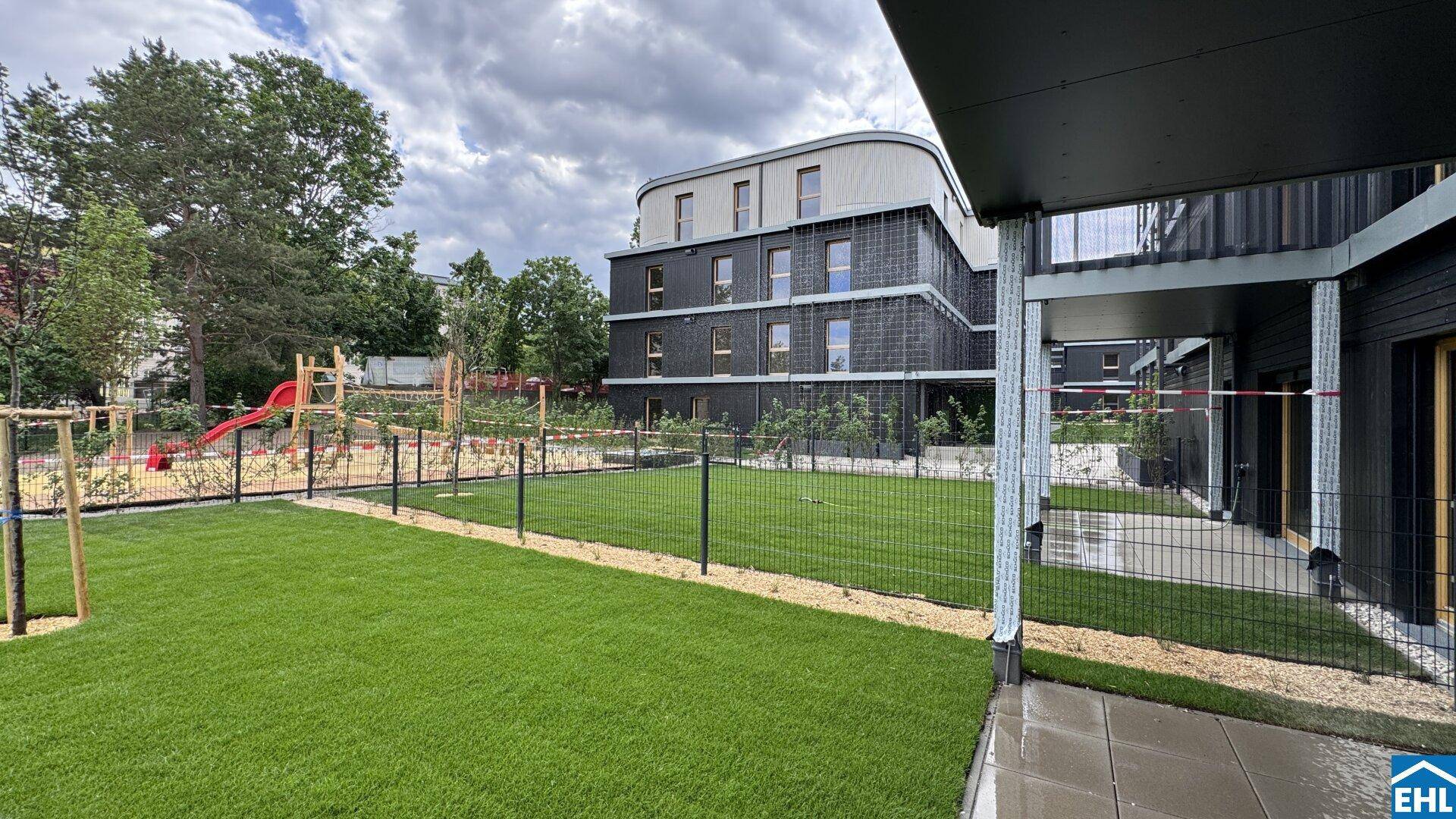 Grüne Rasenfläche neben einem modernen Gebäude mit Spielplatz im Hintergrund unter bewölktem Himmel.