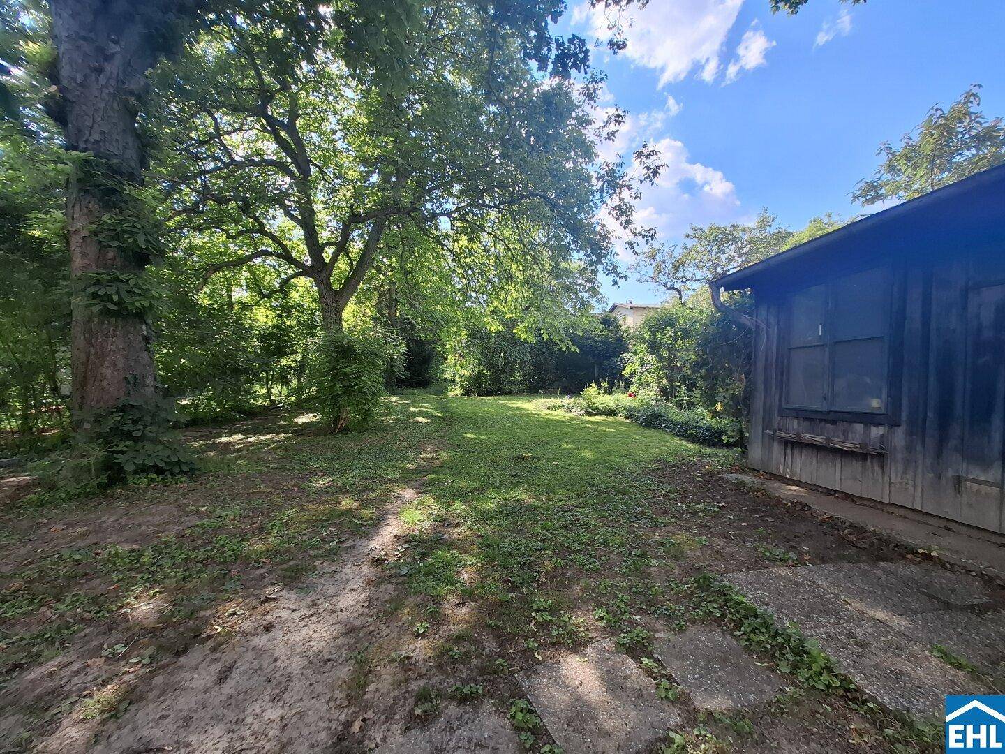 Weitläufiger Gartenbereich mit altem Baumbestand und einer kleinen Holzhütte unter blauem Himmel.