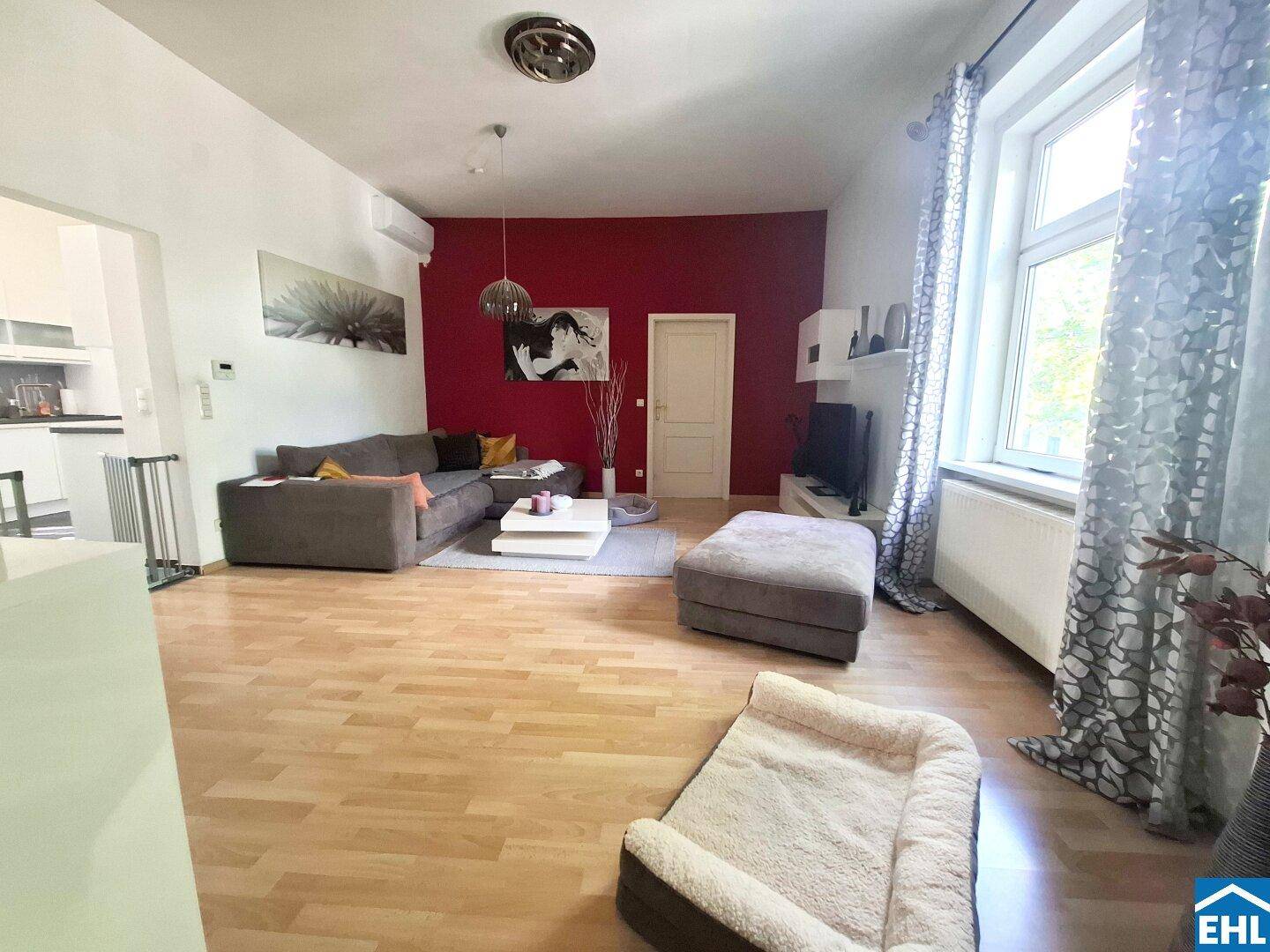 Geräumiges Wohnzimmer mit Holzboden, roter Akzentwand und großem Fenster für viel Licht.