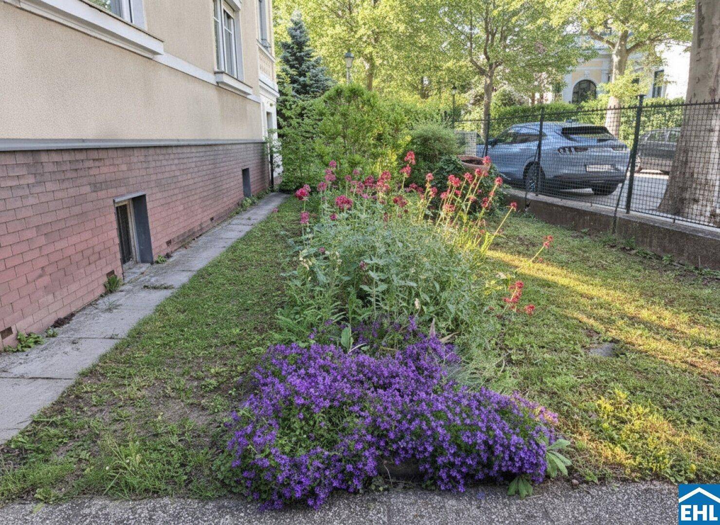 Schmaler Vorgartenstreifen entlang des Gebäudes mit bunten Blumenbeeten und einem Zaun.