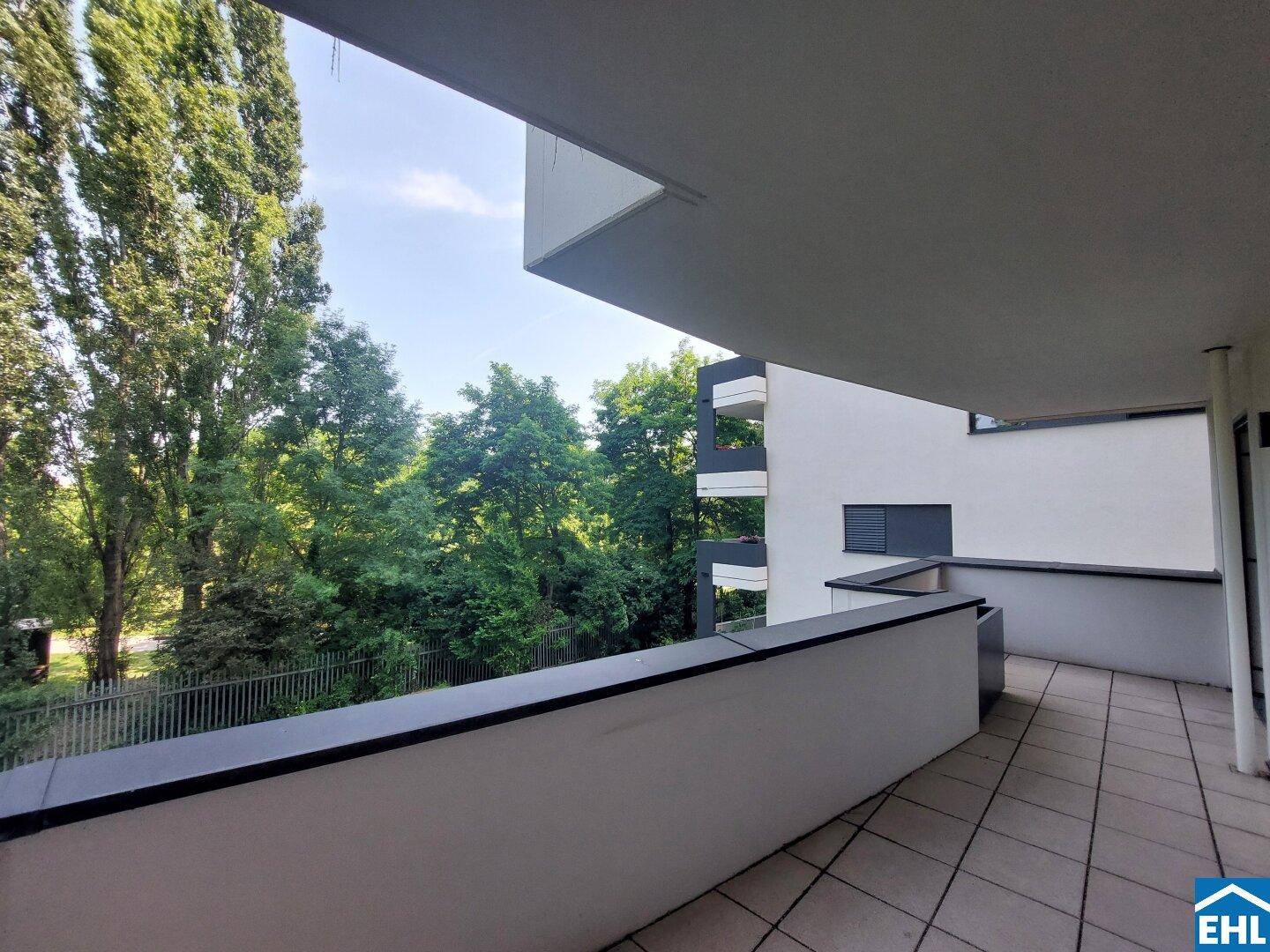 Heller Balkon mit Fliesenboden und Blick auf die umliegende Grünfläche sowie angrenzende Gebäude.