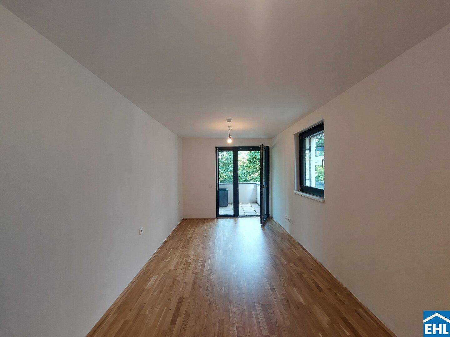Heller Wohnraum mit Parkettboden, weißen Wänden und Zugang zu einem kleinen Balkon mit Grünblick.