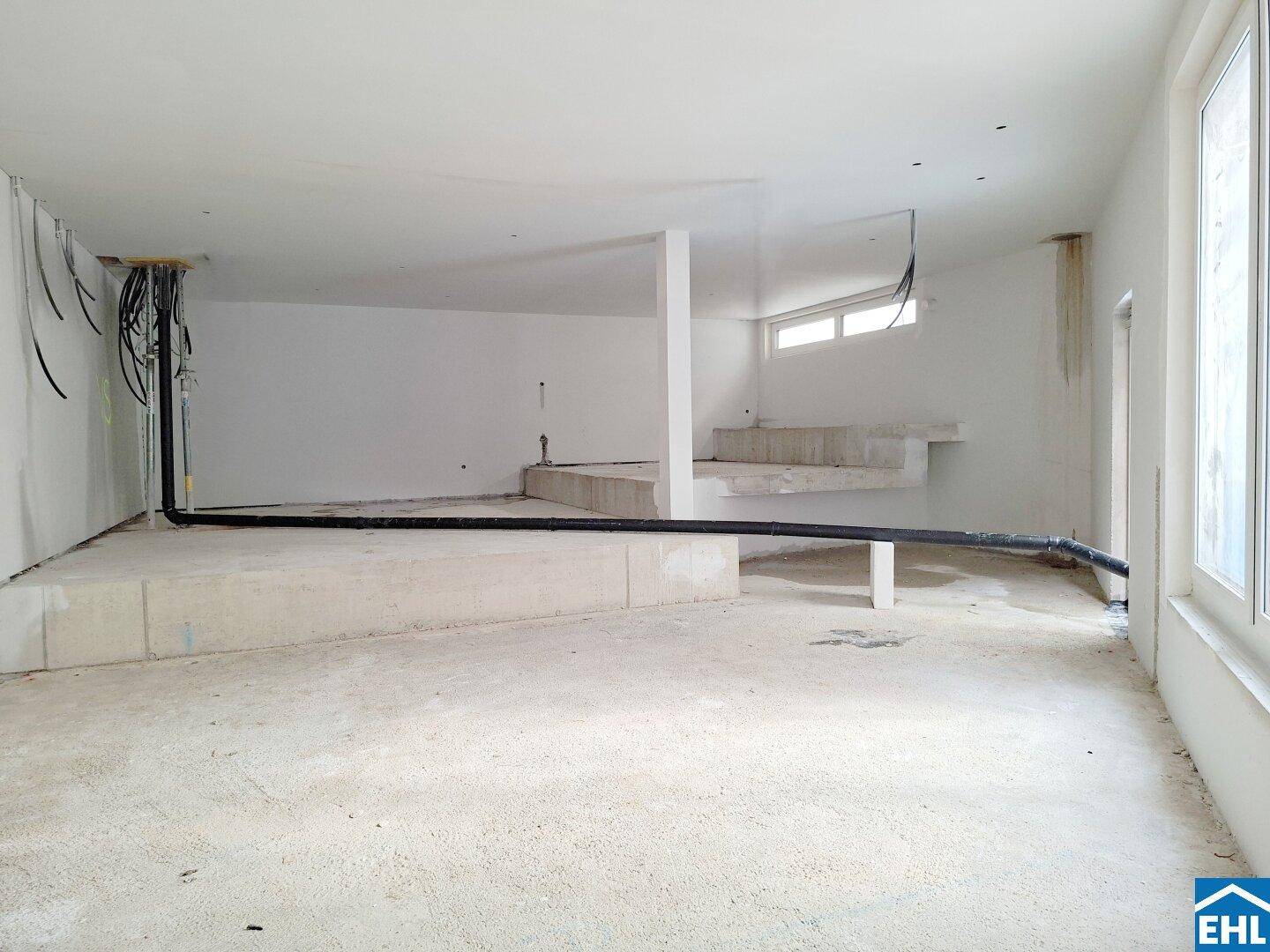 Blick in einen unfertigen Raum mit Betonstufen, sichtbaren Installationen und großen Fenstern.