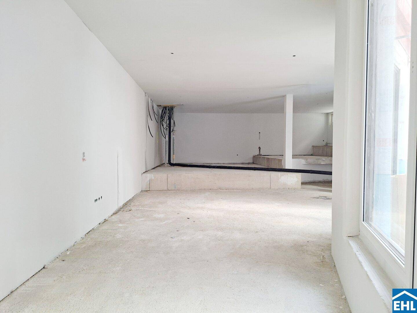 Unfertiger Innenraum mit sichtbaren Kabeln und Rohren, weißen Wänden und einem Betonboden.