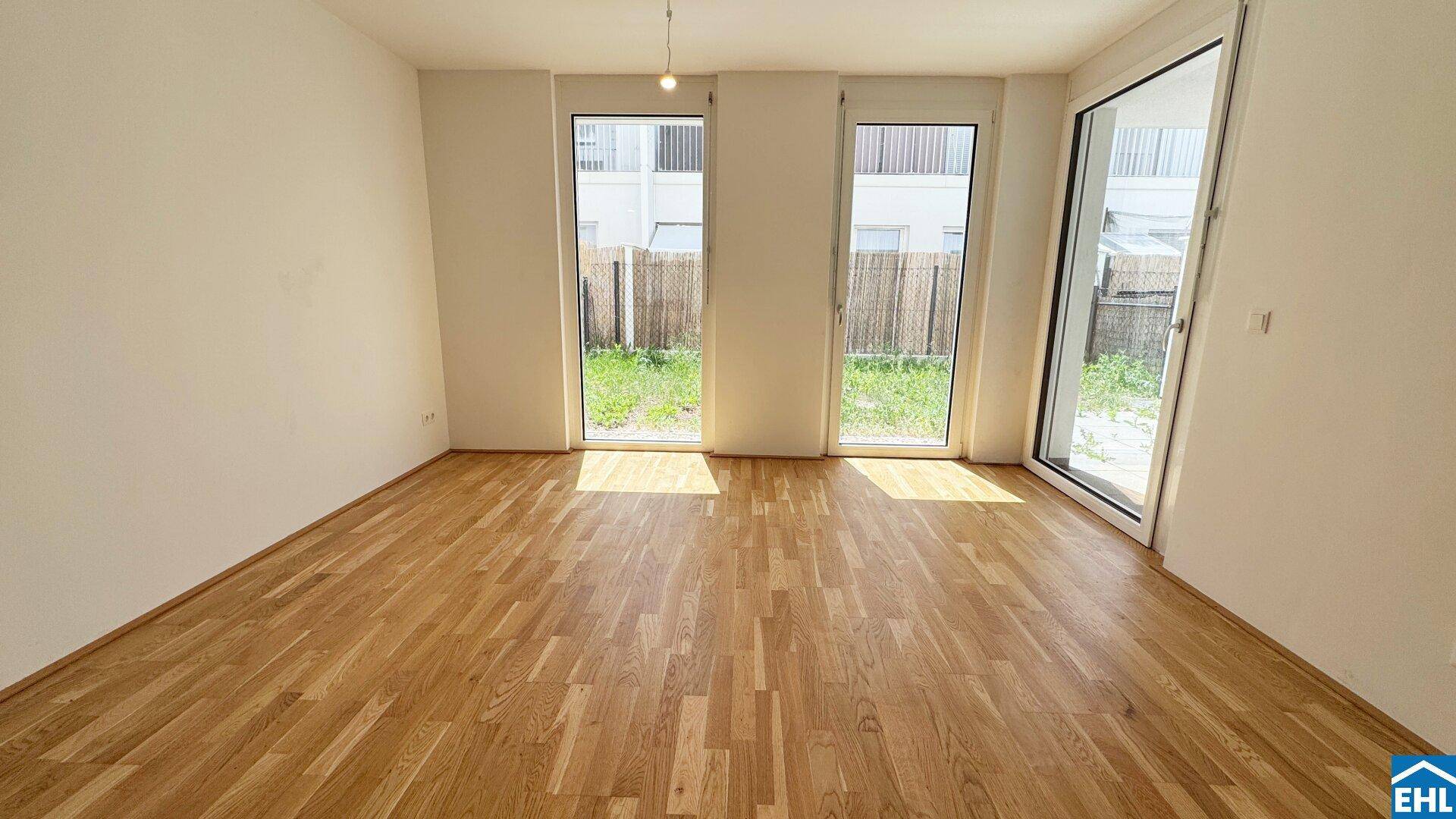 Heller, leerer Raum mit Holzboden und großen Fenstertüren, die Zugang zum Garten bieten.