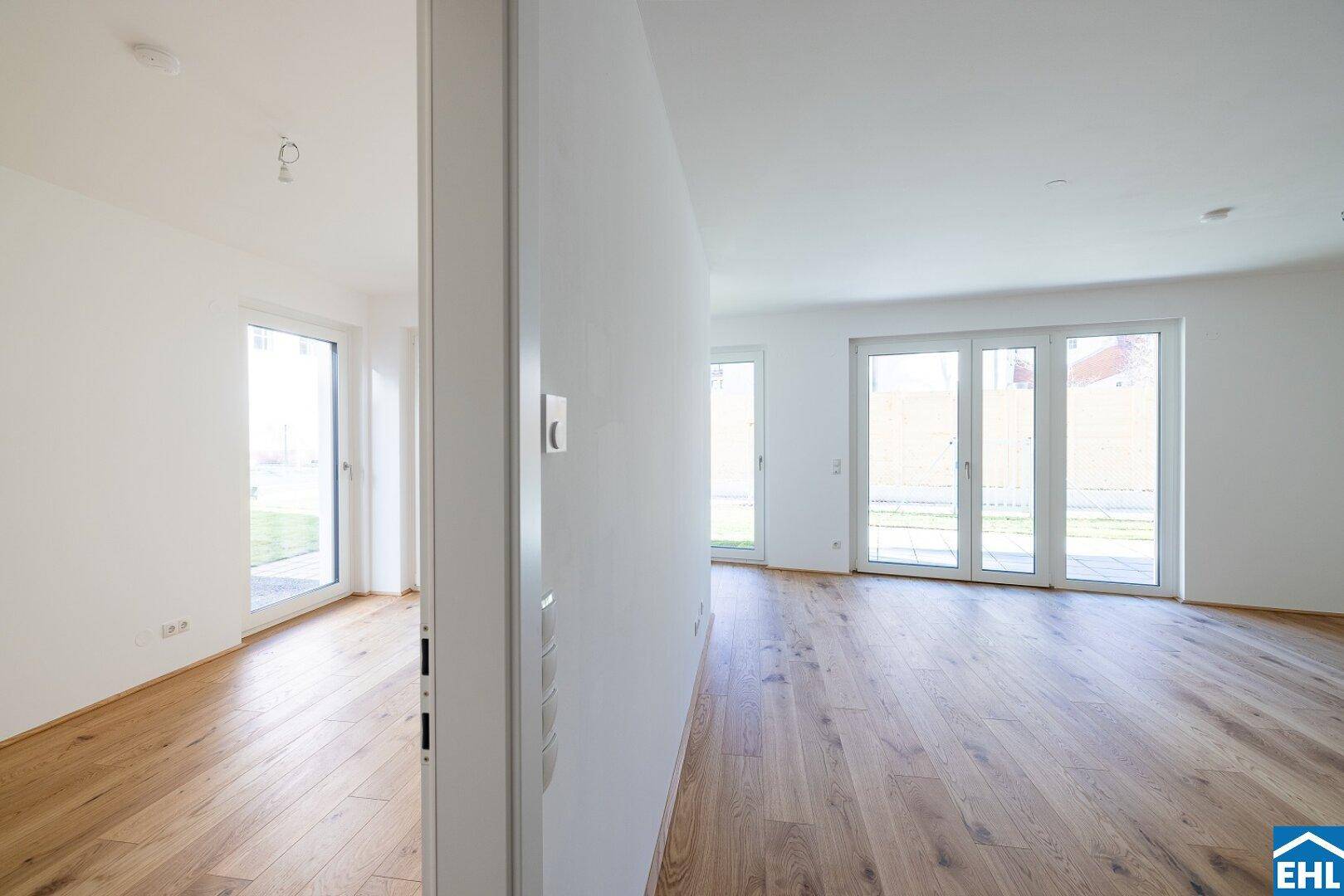 Blick durch eine Tür in einen hellen, leeren Raum mit Holzboden und großen Fenstern.