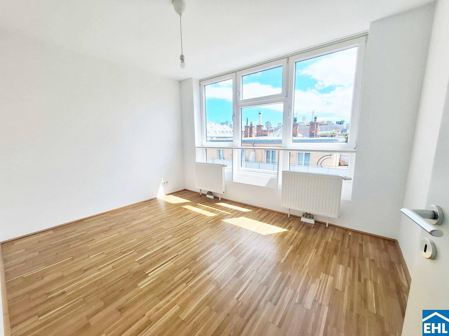 Helles Zimmer mit Parkettboden und großen Fenstern, die viel Tageslicht hereinlassen.